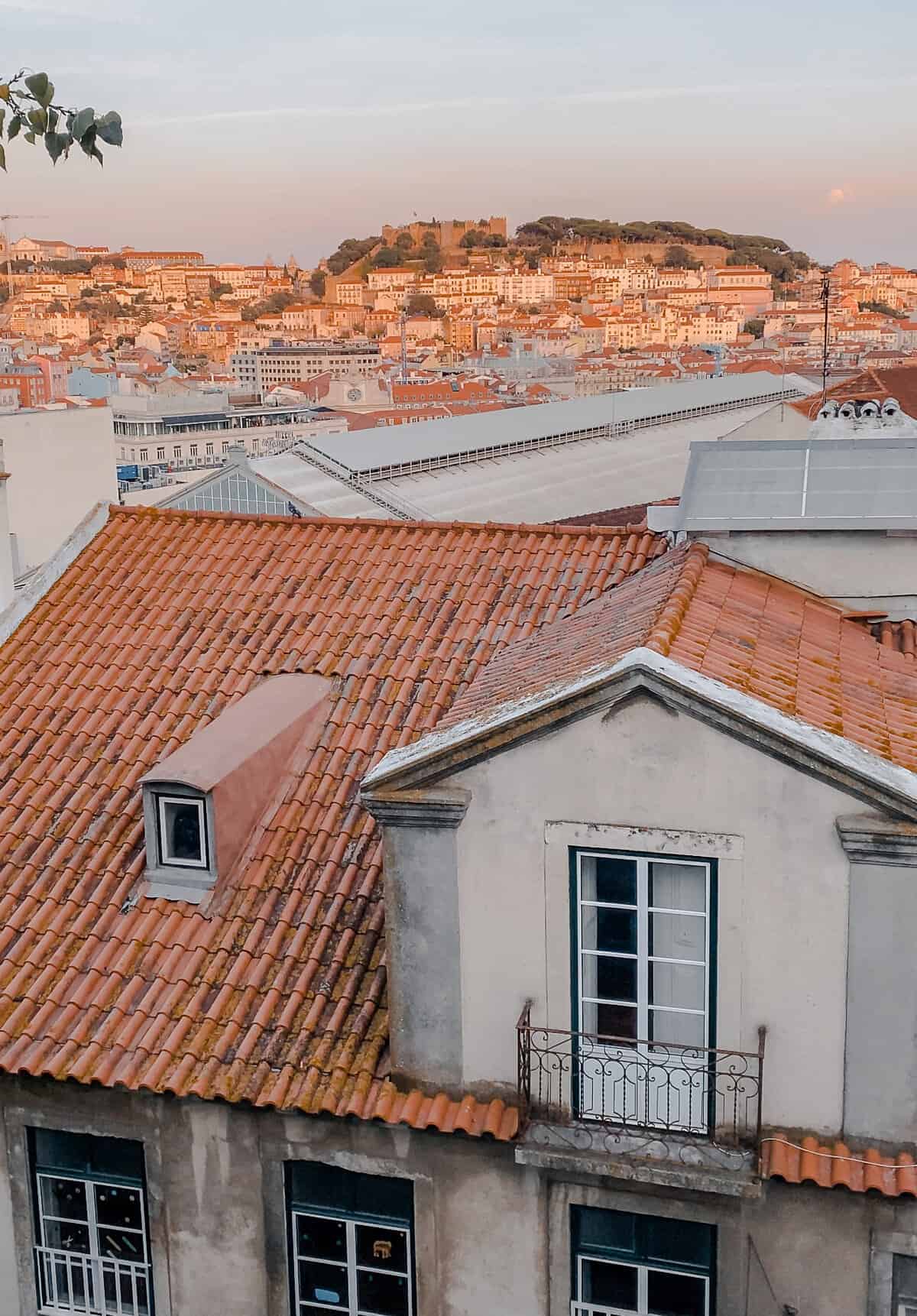 Rooftop view of Lisbon at sunset. The sky is turning pink and the white buildings have an orange glow.