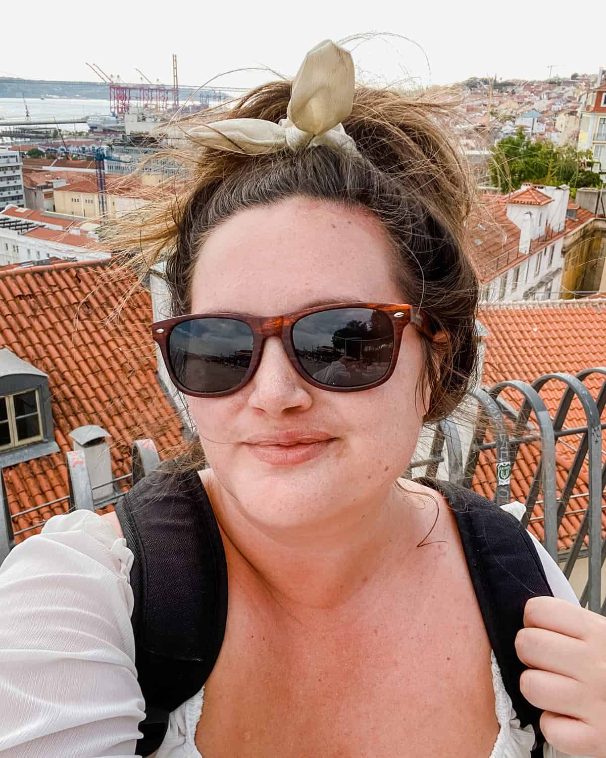 Solo female traveler taking a selfie from one of Lisbon's famous miradouros. Construction, rooftops and Taugus River in background.