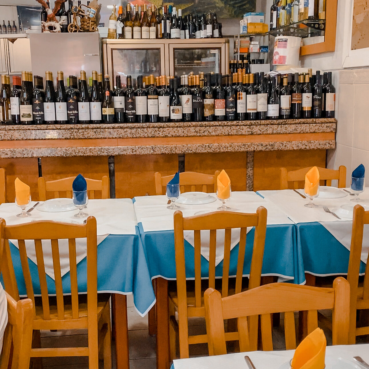 A typical Portuguese restaurant. The counter is lined with Portuguese wine. There are blue table clohts and yellow napkins.